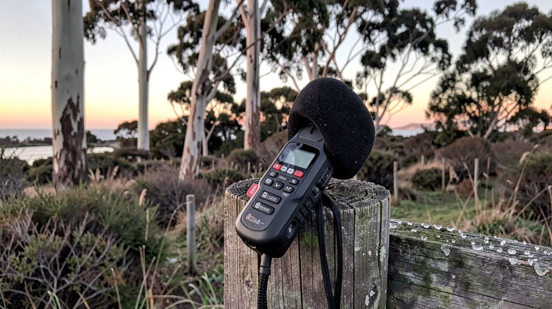 A handheld audio recorder with a foam windscreen rests on a weathered fence post at dawn with coastal bush behind